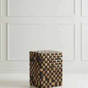 Könnte beinhalten: Ein quadratischer Beistelltisch mit einem geometrischen Muster aus abwechselnd schwarzen und beigefarbenen Quadraten. Der Tisch besteht aus einem Material, das Holz oder ein Holzverbundstoff zu sein scheint. Der Tisch hat eine Würfelform und steht vor einer weißen Wand.