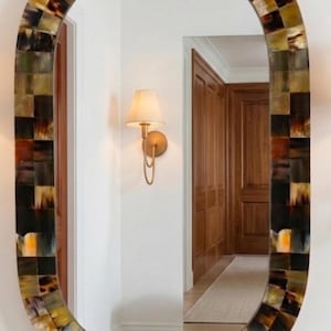 Puede incluir: Un espejo de pared ovalado con un marco ancho compuesto por pequeños azulejos cuadrados multicolores en tonos marrón, negro, bronceado y crema. La superficie reflectante muestra un pasillo con una lámpara de pared y puertas de madera.