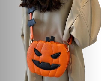 Może przedstawiać: Pomarańczowa torebka na ramię w kształcie dyni z czarną twarzą jack-o'-lantern, trójkątnymi oczami i wyszczerbionymi ustami. Torebka posiada pasujący pomarańczowy pasek ozdobiony małymi czarnymi akcentami w kształcie nietoperzy oraz napisem ELVORA.