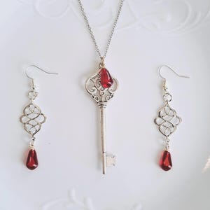 May include: A silver-toned key pendant necklace with a red teardrop-shaped gem, flanked by matching earrings. The earrings and pendant feature an ornate, openwork design. The jewelry set is displayed on a white surface.