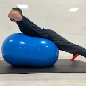 Puede incluir: Una persona se ejercita sobre una gran pelota de ejercicios azul brillante. La persona lleva una camiseta negra, pantalones negros y calcetines naranjas. La pelota de ejercicios está sobre una esterilla negra.
