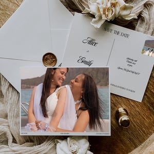 May include: A white Save the Date card featuring a photograph of two people embracing on a boat, with the text Alice and Emily, Saturday 26th October 2024, and Queen, New York City, USA. Includes a white envelope with a gold wax seal and white flowers.