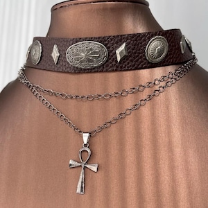 Könnte beinhalten: Ein braunes Leder-Choker-Halsband mit silberfarbenen Verzierungen, darunter runde und rautenförmige Nieten, und einem ovalen Mittelstück. Eine silberne Kette mit einem Ankh-Anhänger hängt darunter.