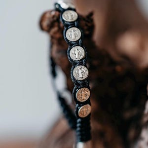 Könnte beinhalten: Schwarz geflochtenes Armband mit silberfarbenen runden Charms. Jeder Charm zeigt eine detaillierte religiöse Figur. Das Armband wird vor einem verschwommenen Holzgrund präsentiert, was das aufwendige Design und die Handwerkskunst des Schmucks hervorhebt.