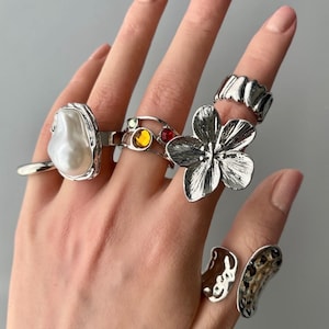 Pode incluir: Uma mão exibindo uma coleção de anéis de prata. Os anéis apresentam vários designs, incluindo uma grande pérola, uma flor e faixas texturizadas. Alguns anéis têm pedras preciosas coloridas.