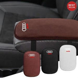 May include: A brown car armrest cover with a white Audi logo is shown, along with three smaller covers in brown, black, and white, each with an Audi logo. The armrest cover is attached to a car seat.