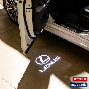 May include: White Lexus car door open, projecting a bright white Lexus logo and text onto the ground. The car's wheel and interior are visible. The door is open, casting a shadow on the concrete surface.