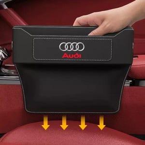 May include: Black car seat organiser with white stitching and the Audi logo in silver and red. The organiser is designed to fit between the car seats and has a hand reaching into it. Yellow arrows point down.