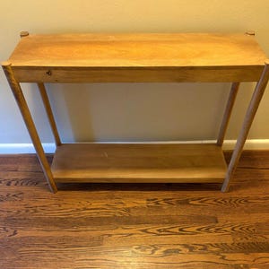 Puede incluir: Una consola de madera de color claro con una parte superior rectangular y un estante inferior. La mesa tiene patas anguladas y esquinas redondeadas. La veta de la madera es visible, dándole un aspecto natural.