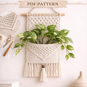 Può includere: Appendiabiti per piante all'uncinetto color crema con una tasca che contiene una pianta verde con variegatura bianca. Il design include una frangia in macramè e un tassello in legno. Il testo recita "CROCHET PLANT HANGER PATTERN PDF PATTERN".