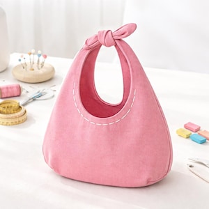 Puede incluir: Un bolso tote de tela rosa con un asa anudada. El bolso tiene una forma redondeada con una abertura ancha y un detalle cosido en blanco. El bolso está sobre una superficie blanca, con útiles de costura en el fondo.