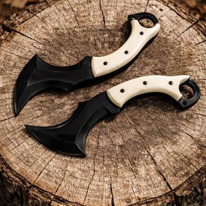 May include: Two black karambit knives with cream-colored handles are displayed on a weathered wooden surface. The blades have a curved design, and the handles feature a finger ring. The knives are designed for tactical or self-defense purposes.