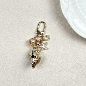 May include: Gold-coloured keychain with a seashell charm, a pearl-centred flower, a starfish, and a letter "G" pendant. The seashell has brown accents. The keychain has a lobster clasp.