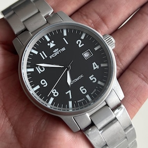 May include: A stainless steel wristwatch with a black face. The watch features white numerals, hands, and a date display at the 3 o'clock position. The watch has the word "FORTIS" printed on the face. The watch is on a stainless steel strap.