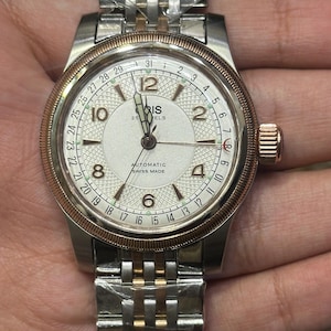 Könnte beinhalten: Eine silber- und goldfarbene Armbanduhr mit weißem Zifferblatt. Die Uhr hat eine goldene Lünette, eine Datumsanzeige und ein silber- und goldfarbenes Armband. Das Zifferblatt zeigt die Marke "ORIS" und die Worte "AUTOMATIC SWISS MADE".