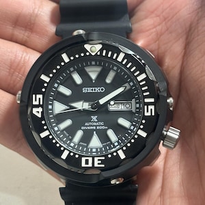 Op de afbeelding: Zwarte Seiko automatische duikershorloge met een zwarte rubberen band. De wijzerplaat is zwart met witte markeringen en wijzers. De lunette heeft witte markeringen op 15, 30 en 45. Het horloge heeft een datumweergave op 3 uur.