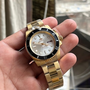 May include: Gold-toned wristwatch with a black bezel and a silver face. The watch features a gold band and a date display. The watch face has the word "ORIENT" printed on it. The watch is held in a hand.