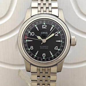 May include: A stainless steel Oris watch with a black dial. The watch features a date display, luminous hands and markers, and a stainless steel bracelet. The dial reads "ORIS 25 JEWELS AUTOMATIC SWISS MADE".