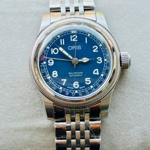 May include: A stainless steel wristwatch with a blue face. The watch has a silver band and a round bezel with a fluted edge. The watch face displays the brand name "ORIS" and the words "BIG CROWN AUTOMATIC" and "SWISS MADE".