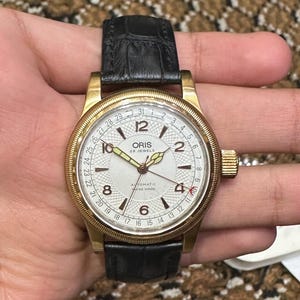 May include: A gold-toned Oris wristwatch with a white face, black leather band, and a date display. The watch features a textured dial, gold hands, and the text "ORIS 25 JEWELS AUTOMATIC SWISS MADE".