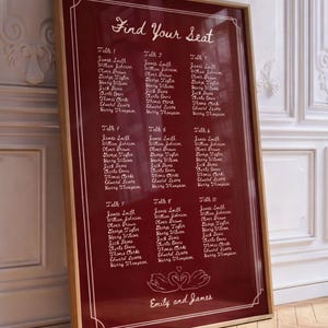 May include: A framed seating plan with a deep red background and white text. The text reads "Find Your Seat" at the top, followed by guest names organised by table. The bottom features two swans and the names "Emily and James".
