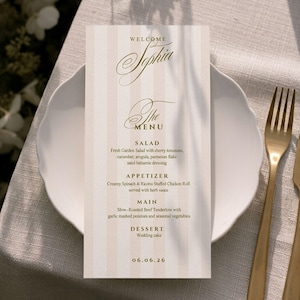 May include: A vertical rectangular menu card with light beige vertical stripes and gold text. The text reads Welcome Sophia, The Menu, Salad, Appetizer, Main, Dessert, and the date 06.06.26. The menu is placed on a white plate with a gold fork.