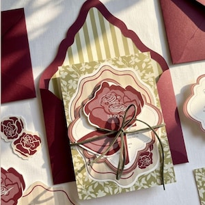 May include: Burgundy and cream wedding invitation suite with floral and striped patterns. The invitation features a large rose design and is tied with a green ribbon. Includes matching envelopes and decorative stickers.