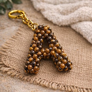 Può includere: Un portachiavi artigianale a forma di lettera "A", realizzato con piccole perline rotonde marroni e nere. Le perline formano la lettera, con una catena e un fermaglio dorati. Il portachiavi è appoggiato su un tessuto beige strutturato.