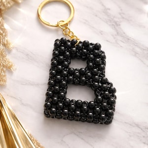 May include: Black beaded letter "B" keyring with a gold-coloured ring. The letter is made of small, round, black beads. The keyring is on a white and grey marble surface.