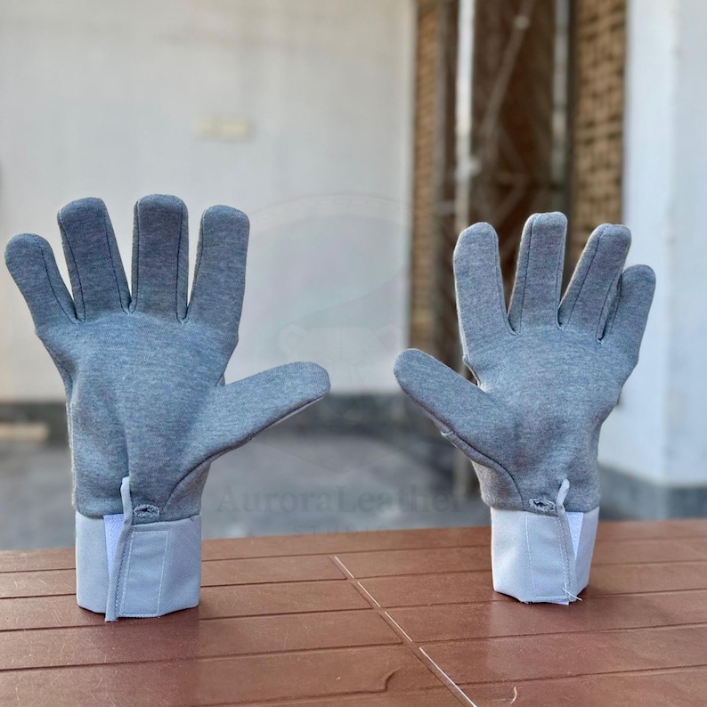 May include: A pair of gray gloves with white cuffs. The gloves are made of a soft material and have a simple design. The cuffs are rectangular and have a small button detail. The gloves are displayed on a brown surface.