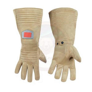 May include: A pair of tan leather gloves with a long gauntlet design. The gloves feature a rectangular patch with red and silver accents on the left glove and a strap with two black buttons on the right glove.