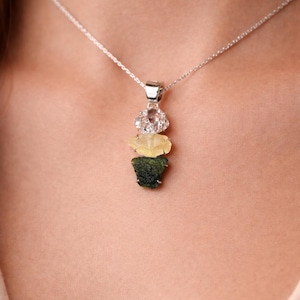 Puede incluir: Un collar de plata con un colgante compuesto por tres piedras preciosas apiladas. La piedra superior es transparente, la del medio es amarilla y la inferior es verde oscuro. El collar se muestra sobre un fondo neutro.