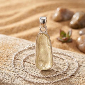 Może przedstawiać: Srebrny naszyjnik z wisiorkiem z jasnożółtym kamieniem szlachetnym. Kamień jest osadzony w srebrze i zawieszony na delikatnym srebrnym łańcuszku. Wisiorek ma gładką, wypolerowaną powierzchnię i nieregularny, wydłużony kształt.