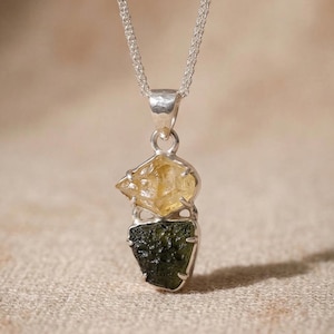 Może przedstawiać: Srebrny naszyjnik z wisiorkiem z surowym kryształem cytrynu nad surowym kryształem mołdawitu. Cytryn jest w kolorze jasnożółtym, a mołdavit w kolorze ciemnozielonym. Wisiorek zawieszony jest na delikatnym srebrnym łańcuszku.
