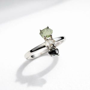 Könnte beinhalten: Ein silberner Ring mit drei ungeschliffenen Edelsteinen in Grün, Klar und Schwarz. Die Edelsteine sind in einer Reihe auf dem Ringband angeordnet. Der Ring wird vor einem weißen Hintergrund präsentiert.