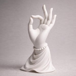 May include: A white ceramic hand sculpture in the 'okay' gesture, adorned with a beaded bracelet. The hand rests on a draped base, creating a serene and artistic display. The sculpture is a decorative item.