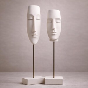 Puede incluir: Dos esculturas de rostros blancos con los ojos cerrados. Cada rostro está montado sobre una fina varilla de metal oscuro y colocado sobre una base rectangular blanquecina. Las esculturas tienen un diseño alargado, ideal para la decoración del hogar. Aproximadamente 30 cm de altura.