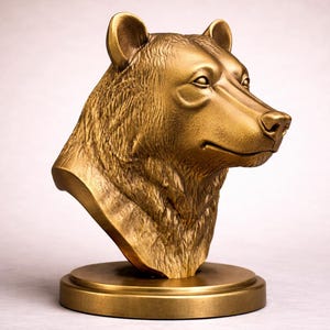 May include: A gold-coloured bear bust sculpture. The bear's head is detailed with fur texture and facial features, mounted on a round base. The sculpture is made of a metallic material, and the overall design is a classic representation of a bear.