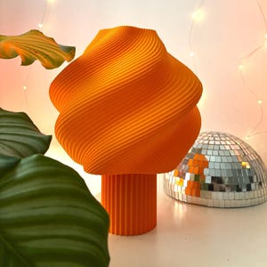 Könnte beinhalten: Eine leuchtend orangefarbene Tischlampe mit einem breiten, spiralförmig gerippten Schirm, der in einen schmaleren, vertikal geriffelten zylindrischen Sockel übergeht. Die Lampe hat eine matte Oberfläche und eine markante geometrische Form.