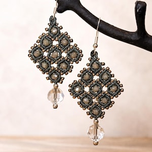 Könnte beinhalten: Ein Paar handgefertigte Makramee-Ohrringe. Jeder Ohrring hat ein rautenförmiges Design in Dunkelgrau mit kleinen, runden, hellgrauen Perlen und goldfarbenen Akzenten. An der Unterseite jedes Ohrrings hängt eine klare, facettierte Kristallperle.