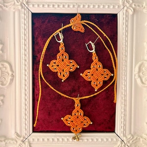 May include: A set of orange macrame jewelry consisting of two earrings and a pendant, each featuring a complex woven knot design. The earrings have silver-toned clasps, and the pendant includes a small gold-toned bee charm at the bottom.