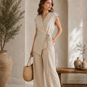 Op de afbeelding: Een beige linnen wikkel top en broek met wijde pijpen. De outfit wordt gecombineerd met een geweven handtas. De afbeelding is gemaakt in een kamer met neutrale tinten met een decoratieve vaas en een houten bank. De outfit is van NooreLuxe Studio.