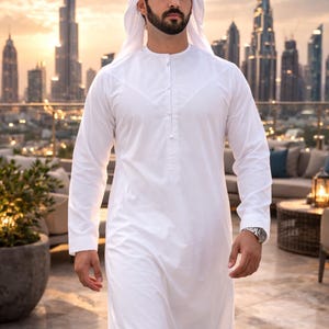May include: A man in a white thobe and traditional headdress walks toward the camera. The long-sleeved garment has a buttoned placket. A watch is visible on the wrist. The background features a cityscape at sunset.