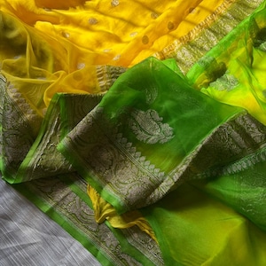 May include: A vibrant yellow and green sari with intricate silver-colored detailing. The sari features a sheer, flowing fabric with a decorative border. The design includes small, circular patterns and a contrasting green section.