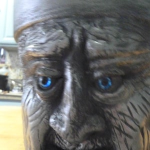 May include: A ceramic container with a sculpted face. The face is gray with blue eyes and a beard. The container has a cylindrical shape and is likely used for storage or decoration.