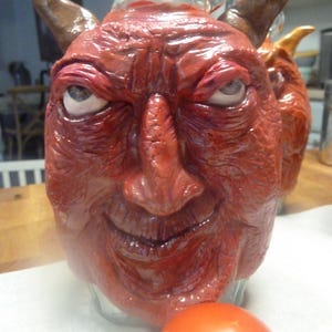 May include: A clear glass jug featuring a sculpted red face with prominent wrinkles, a pointed nose, and two brown horns protruding from the top. The face has white eyes with dark pupils and a subtle smirk. A smooth, round orange tomato sits in the foreground.