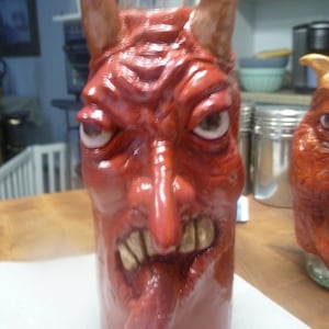 May include: A glass bottle featuring a sculpted, textured red devil face with prominent horns, wide eyes, a long nose, and a mouth with visible teeth and a protruding tongue. The surface has a glossy finish, and the neck of the bottle is clear glass.