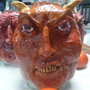 May include: A clear glass bottle featuring a sculpted, textured, reddish-brown face with prominent horns, deep-set eyes, and visible teeth. The sculpture covers the front of the bottle, displaying a detailed, wrinkled skin texture and a stern expression.