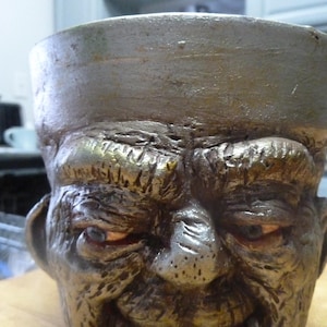 May include: A ceramic planter shaped like a face with a textured, aged look. The face has a furrowed brow, deep-set eyes, and a wide, toothy grin. The planter is painted in shades of brown and gray.
