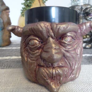 May include: A handcrafted, decorative container with a goblin face design. The container is painted in shades of bronze and purple, with a black interior. The goblin face features detailed wrinkles, a prominent nose, and pointed ears. A unique piece for home decor.